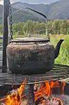 Kaffekjel over flammer på en grillrist, med grønne trær og fjell i bakgrunnen.