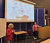 To personer i røde klær står i et møterom foran en illustrert presentasjon.