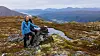 To turgåere sitter ved en bekk i et steinete fjellandskap med fjell i bakgrunnen.