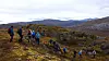 Fjellvandrere går på rekke gjennom et steinete høstlandskap med fjell i bakgrunnen.