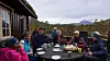 Gruppe voksne rundt et bord utenfor en hytte, med fjell og skog i bakgrunnen.