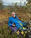 Person i blå jakke sitter blant lyng og busker i et høstlig fjellandskap.
