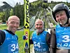 Tre skihoppere i blå drakter foran en skihoppbakke.