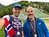 To menn med medaljer foran en grønn hoppbakke i fjellandskap.