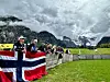 Mennesker med norske flagg ved et gjerde foran grønne marker og fjell.