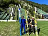 Tre personer med ski foran en skihoppbakke omgitt av skog og gress.