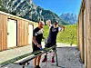 To personer med sportsutstyr ved trebygninger i et grønt fjellandskap.