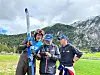Tre skiutøvere står sammen på en gressbane foran fjell.