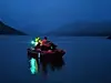 Liten opplyst båt med personer på mørkt, stille vann mellom fjell.