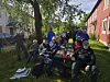 Gruppe mennesker samlet rundt et bord i en grønn hage mellom trehus.