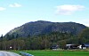 Skogkledd fjell, hus og vei i et grønt bygdelandskap under blå himmel.