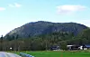 Skogkledd fjell, hus og vei i et grønt bygdelandskap under blå himmel.