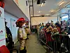 Brannmannskledd figur foran en folkemengde bak sperringer innendørs.