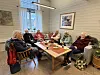 En gruppe voksne sitter rundt et bord med kaffe og mat i et lyst rom.