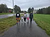 Voksne og barn går sammen på en våt vei gjennom et grønt bygdelandskap.