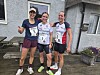 Tre kvinnelige løpere med medaljer og startnumre poserer foran et hus.