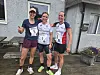 Tre kvinnelige løpere med medaljer og startnumre poserer foran et hus.