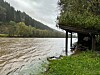 Oversvømt gapahuk ved ei flomstor elv med skogkledde åser i bakgrunnen.