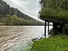 Oversvømt gapahuk ved ei flomstor elv med skogkledde åser i bakgrunnen.