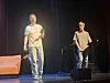 To scenekunstnere på en belyst scene med sofa, høyttaler og mørke scenetepper.