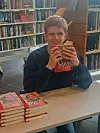 Smilende ung mann holder en bok ved et bord i et bibliotek.