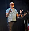 Foredragsholder med briller gestikulerer på en mørklagt scene.