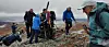 Turgåere med turutstyr på steinete kystterreng i overskyet vær.