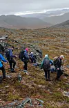 Turgåere med sekker går gjennom et steinete fjellandskap under en overskyet himmel.