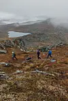 Tre turgåere går i et steinete fjellandskap med vann og tåke i bakgrunnen.