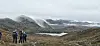 Vandrere i et karrig landskap med dampende fjell og snødekte høyder i bakgrunnen.
