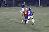 Fotballspillere i røde og blå drakter kjemper om ballen på en kunstgressbane.