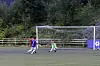 To fotballspillere ved et mål på en utendørsbane foran et skogkledd område.