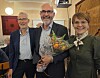 Tre personer smiler mot kameraet i et innendørslokale; mannen i midten holder blomster og en gave.