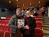 To menn står i en kinosal og holder fram en bok foran røde stoler.
