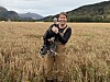 Smilende dame med en kalkun i en kornåker foran skog og fjell.