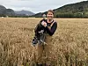Smilende dame med en kalkun i en kornåker foran skog og fjell.