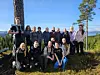 En gruppe mennesker poserer utendørs ved trær med fjell og innsjø i bakgrunnen.