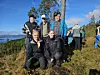 Ei gruppe turgåere poserer på en fjellkledd høyde med utsikt over vann og fjell.