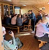 Mange turkledde mennesker sitter samlet rundt langbord i et lyst rom med trevegger og tregulv.
