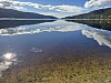 Stille innsjø med speilblankt vann, fjell, skyer og steinete bunn i forgrunnen.