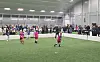 Barn spiller fotball på kunstgress i en stor innendørshall med tilskuere langs sidelinjen.