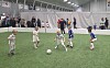Barn spiller fotball på kunstgress innendørs, med tilskuere bak en avskjerming.