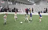 Barn spiller fotball på kunstgress innendørs, med tilskuere bak en avskjerming.