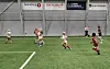 Barn spiller fotball på kunstgress i en innendørshall med sponsorlogoer på veggen.
