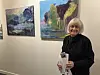 Kvinne med brosjyre og papir foran to fargerike malerier i et galleri.