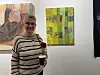 Smilende kvinne med briller og stripete genser, med kaffekopp og en rose foran to kunstverk på en hvit gallerivegg.