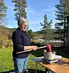 Kvinne ved et utebord med gryte og rødt tekstilmateriale i en solrik hage ved vannet.