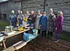 Gruppe voksne står ved et utebord med mat og kjøkkenutstyr foran en gammel trebygning.