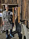 Person i mønstret forkle foran en gammel trebygning med trestang.