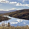 Stille innsjø med speilblank fjellrefleksjon, skogkledde åser og skyer på blå himmel.